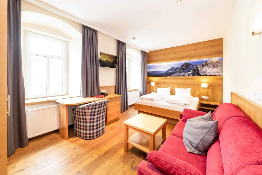 Bright hotel room with a double bed, desk, red sofa, and mountain views